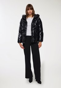 Giacca a vento nera con finitura lucida, cappuccio, vita aderente e maniche lunghe, indossata sopra un top bianco con piccoli embellishments.