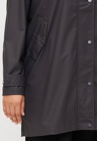Close-up of a person's hand holding the hem of a black waterproof jacket, featuring snap buttons and a side pocket.