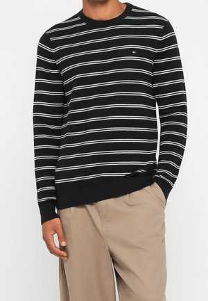 Sort sweater med hvide horisontale striber båret af en person med beige bukser, højre hånd i lommen, stående mod en ensfarvet baggrund.
