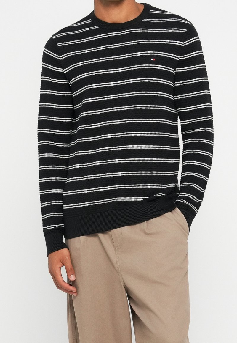 Pull noir à rayures horizontales blanches porté par une personne avec un pantalon beige, main droite dans la poche, debout devant un fond uni.