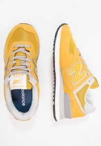 Ett par gula och grå New Balance 574 sneakers, en som visar ovanifrån med snörena, den andra som visar sidan med logotyp och texturerad sula.