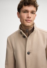 Beige wolmix jas met een hoge kraag, voorzien van een bruin, met rits afsluitbaar binnenlaag en twee zwarte knopen voor sluiting. Soepele textuur.