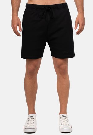 Mężczyzna w czarnych spodenkach ze sznurkiem i białych niskich trampkach, stojący na jednolitym białym tle.