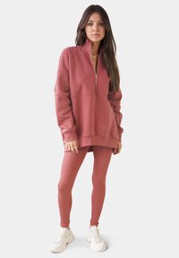 Felpa rosa chiaro con zip e colletto alto, abbinata a leggins aderenti coordinati e sneakers bianche, realizzata in morbido tessuto e con cuciture minime.