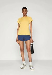 Geltonos spalvos trumpomis rankovėmis marškinėliai, mėlyni šortai, balti kojinės ir sportbačiai. Modelis laiko juodą vandens butelį; minimalistinis dizainas, laisvas stilius.