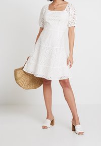 Robe blanche à manches courtes avec encolure carrée, motifs en œillets et jupe à volants. Associée à un sac en paille tissé et des mules à talons blocs blancs.