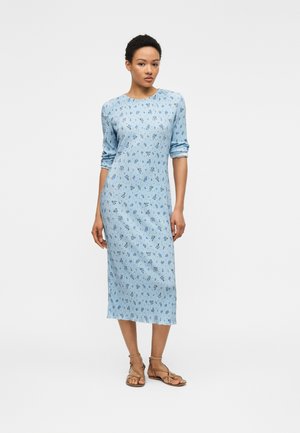 Femeie în picioare purtând o rochie midi cu mâneci lungi, de un albastru deschis, cu imprimeu floral mic și sandale maro joase.