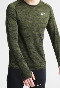 Långärmad träningsshirt i mörk olivgrön med texturerat tyg, med rund halsringning och Nike-logotyp på bröstet.