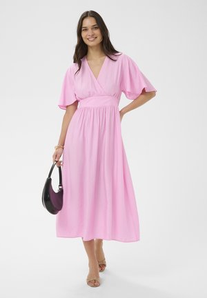 Mujer con un vestido midi rosa de mangas anchas sosteniendo un bolso negro y morado, sonriendo y posando con una mano en la cadera.