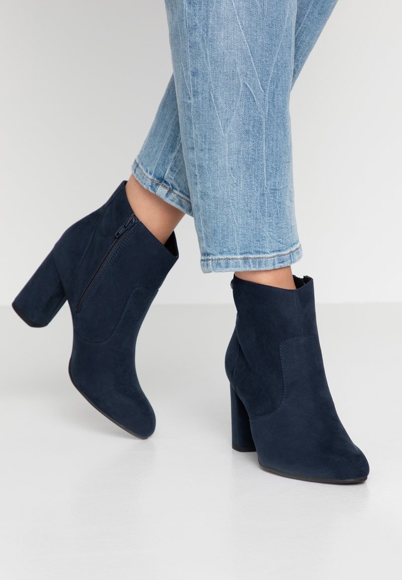 Personne portant des bottines en daim bleu avec talons blocs et jeans courts bleu clair, debout sur un sol blanc lisse.