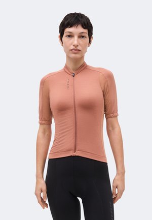 Femme aux cheveux courts et foncés portant un maillot de cyclisme rose saumon ajusté et un cuissard noir, se tenant devant un fond uni.
