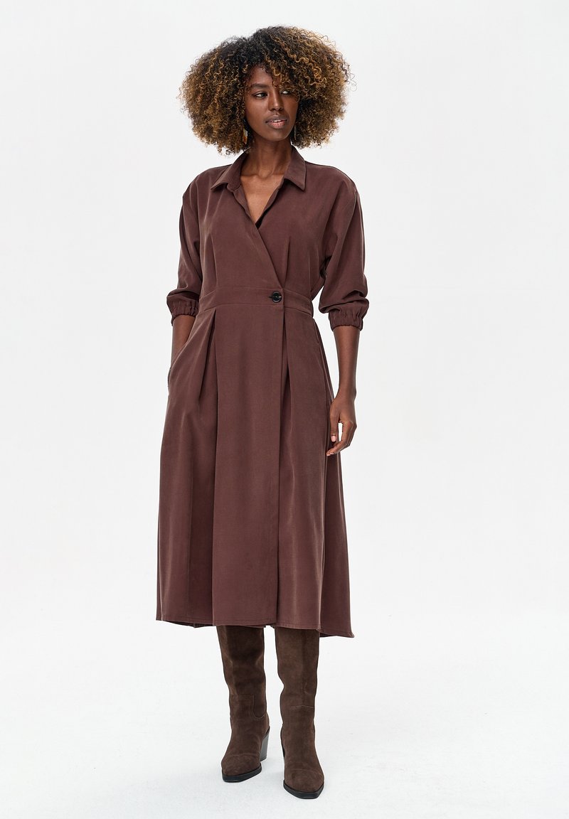 Brązowa, długorękawowa sukienka kopertowa z kołnierzem i zapięciem na guziki. Charakteryzuje się pionowymi plisami i gładką fakturą, zestawiona z brązowymi kozakami za kolano.