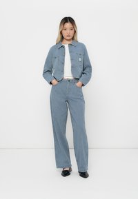 Vrouw staat met handen in de zakken, gekleed in een blauw gestreepte jacket en bijpassende wijdlopende broek over een wit topje en zwarte schoenen.