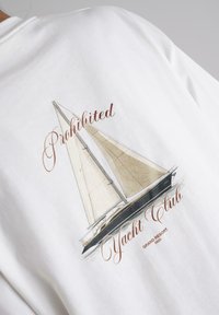 White cotton t-shirt featuring a graphic of a sailboat with beige sails, text "Prohibited Yacht Club" in brown, and "Grand Resort" below.