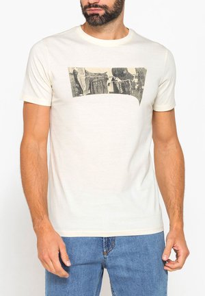 Uomo con la barba che indossa una maglietta crema a maniche corte con una grafica rettangolare bianca e nera sul petto e jeans blu.