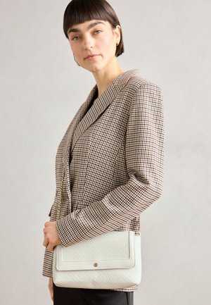 Balta siuvinėta petinė rankinė su lygia apdaila, turinti užsegimą ir subtilią aparatūrą, laikoma asmens, vilkinčio patikrintos raštą švarką.