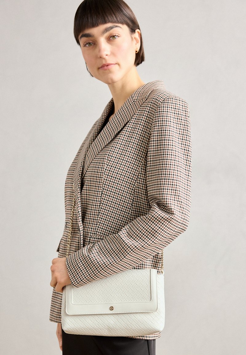 Bolso de hombro blanco acolchado con un acabado suave, que presenta una tapa y un hardware sutil, sostenido por una persona que lleva un blazer a cuadros.