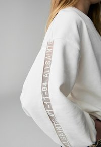 Vit sweatshirt med ribbad textur, med en logoband i grått på ärmen. Designen betonar en avslappnad passform och upptäcktsstil.