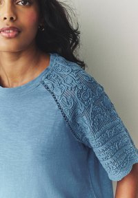 Light blue T-shirt with intricate lace detail on the short sleeves, featuring a soft texture and a round neckline.