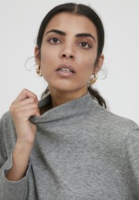 Pulover gri tricotat cu guler înalt, având o textură moale și mâneci largi. Cercei rotunzi aurii pun în evidență aspectul.