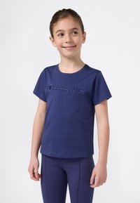 Niña sonriente, vestida con una camiseta y leggings oscuros de Champion que combinan, de pie contra un fondo claro y liso.
