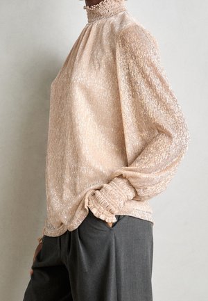 Osoba ubrana w błyszczącą beżową bluzkę z wysokim kołnierzem i marszczonymi długimi rękawami oraz węglowoszare spodnie, stojąca z jedną ręką w kieszeni.