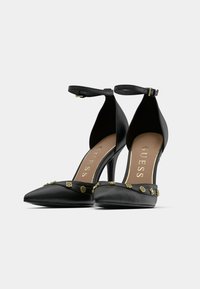 Crne cipele na visoku petu s remenčićem oko gležnja, špicastim vrhom, glatkom teksturom kože i ukrasnim zlatnim detaljima na prednjem dijelu.