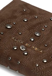 Brązowy portfel z zamszu, ozdobiony przeszyciami, metalowymi ćwiekami i kamieniami, z wytłoczonym złotym logo marki "ALMA EN PENA."