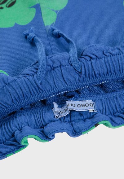 Ceinture en tissu bleu avec un élastique froncé et une bordure verte, comportant une étiquette cousue dans la couture. Texture lisse sans motifs visibles.