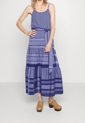 Frau trägt ein ärmelloses blau gemustertes Maxikleid mit Taillengürtel und hellbraunen Clogs mit Absatz, steht vor einem schlichten Hintergrund.