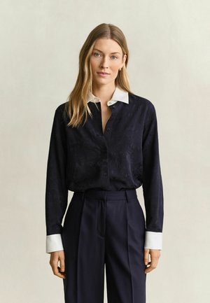 Femme aux cheveux blonds portant un chemisier noir boutonné avec col et poignets blancs, et un pantalon noir taille haute, debout devant un fond uni.