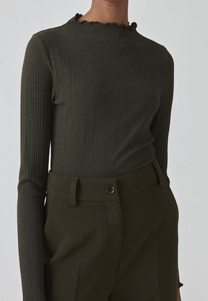 Femme portant un haut à manches longues côtelé vert olive avec un col volanté et un pantalon taille haute vert olive avec fermeture à boutons.