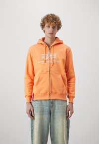 Narancssárga zipzáros kapucnis pulóver, puha anyagból készült, elülső logóval és kenguru zsebbel. Világoskék farmerrel párosítva.
