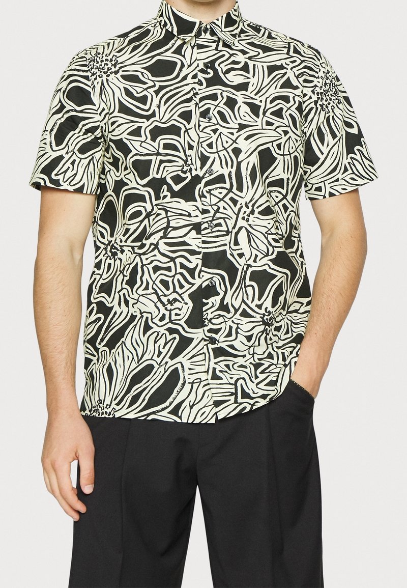 Homme portant une chemise à manches courtes noire et blanche avec un motif floral abstrait et un pantalon noir, une main dans la poche.
