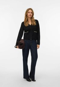 Črni pleteni kardigan z V-izrezom in zapiranjem na gumbe, z dvema sprednjima žepoma s kremnimi obrobami. Kombiniran s temnimi denim kavbojkami in rjavo torbo.
