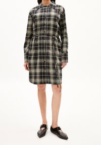 Vestito camicia a quadri nero e verde con maniche lunghe, apertura con bottoni, vita affusolata e dettagli con cordoncino. Abbinato a scarpe basse nere.