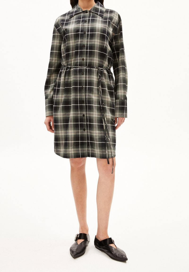 Vestito camicia a quadri nero e verde con maniche lunghe, apertura con bottoni, vita affusolata e dettagli con cordoncino. Abbinato a scarpe basse nere.