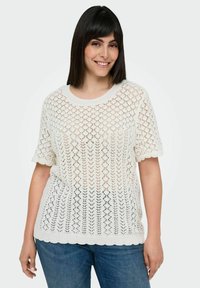 Hvid strikket sweater med diamant hulmønster, korte ærmer og bølget kant, parret med blå jeans.