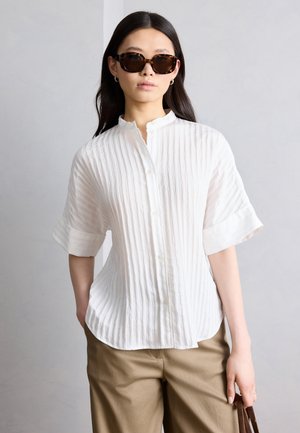 Žena s okuliarmi s vzorom korytnačej mušle, v bielej textúrovanej košeli s krátkym rukávom na gombíky, béžových nohaviciach, stojaca pred sivým pozadím.