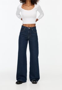 Mujer vestida con jeans oscuros de pierna ancha, top corto de manga larga blanca y zapatos negros, de pie con las manos en las caderas frente a un fondo blanco.
