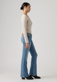 Γυναίκα σε προφίλ, φοράει μπεζ μακρυμάνικη μπλούζα, μπλε καμπάνα τζιν, μαύρες μπότες και κρίκους σκουλαρίκια, με φόντο μια μονόχρωμη γκρι επιφάνεια.