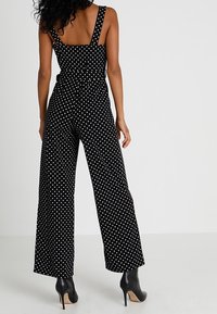 Svart vid byxdress med vita prickar, försedd med tjocka axelremmar och rak rygg, parat med svarta klackstövlar.