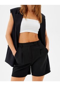 Černé elegantní kraťasy s sklady a bočními kapsami, spárované s bílým vroubkovaným crop topem a černým bezrukávovým blejzrem.