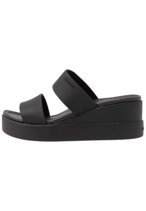 Sandales à talons compensés noires avec deux larges brides. Fabriquées en un matériau lisse ressemblant au caoutchouc, avec une semelle intérieure texturée et une semelle épaisse.