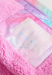 Fleece deken met een roze, fluffy buitenkant en een pastelgradiënt binnenlaag. Waslabel met iconen voor waterdichte en winddichte eigenschappen.