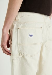 Cream-colored Lee workwear pants feature dual back pockets, contrasting stitching, a branded label, and a relaxed fit.