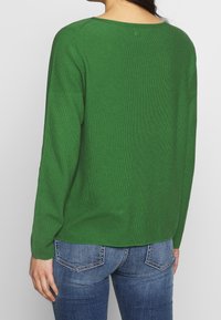 Green, ribbed knit sweater with long sleeves and a wide neckline. Soft texture; hemline sits above the hip, paired with blue denim.