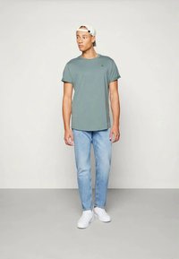 Light green cotton t-shirt with short sleeves, paired with light blue jeans and white sneakers, worn by a model in a neutral background.