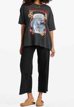 Kobieta ubrana w oversize czarny t-shirt Billabong z grafiką szkieletu surfującego, czarne spodnie jeansowe o skróconych nogawkach z surowymi brzegami oraz jasnobrązowe buty wsuwane.
