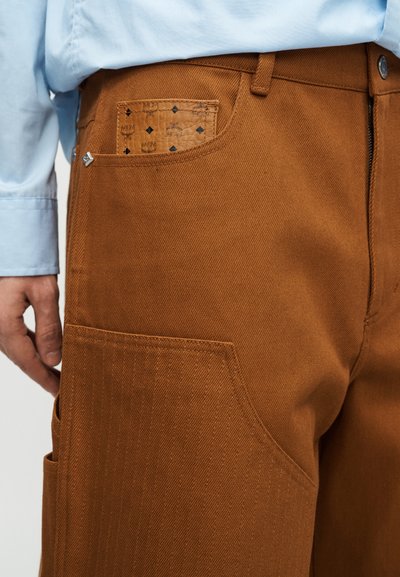 Κοντινό πλάνο ενός ατόμου που φοράει παντελόνια σε σκουρόχρωμο πορτοκαλί με δερμάτινο μοτίβο στο μπροστινό τσέπη και ορατό μανίκι γαλάζιου πουκαμίσου.